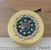 Decorative round compact with gold and gemstone design on a wooden surface, featuring Puca brand.
