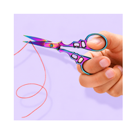 Hand holding Chroma Series rainbow titanium-coated embroidery scissors while trimming fine pink thread against a lavender background