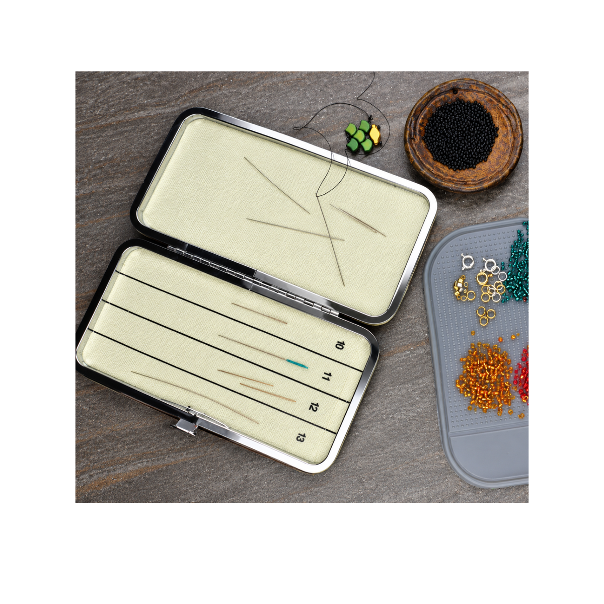 Open double-sided magnetic needle case holding beading needles on a jewellery-making workspace with seed beads, thread and jewellery findings