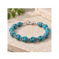 A turquoise and silver beaded bracelet displayed on a textured beige stone surface with a blurred green plant in the background.