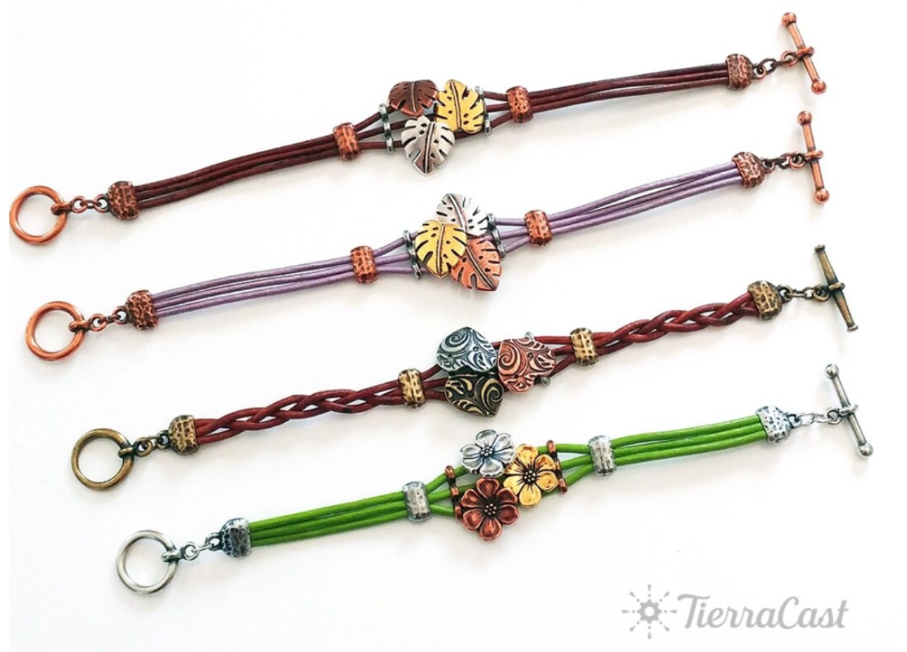 Four braided bracelets in brown, purple, red, and green with floral and leaf charms on a white background.
