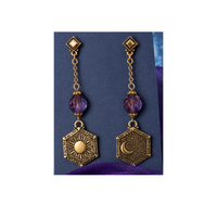 A pair of gold-toned dangle earrings with faceted purple beads and hexagonal charms, one featuring a sun design and the other a crescent moon, displayed on a dark blue card.