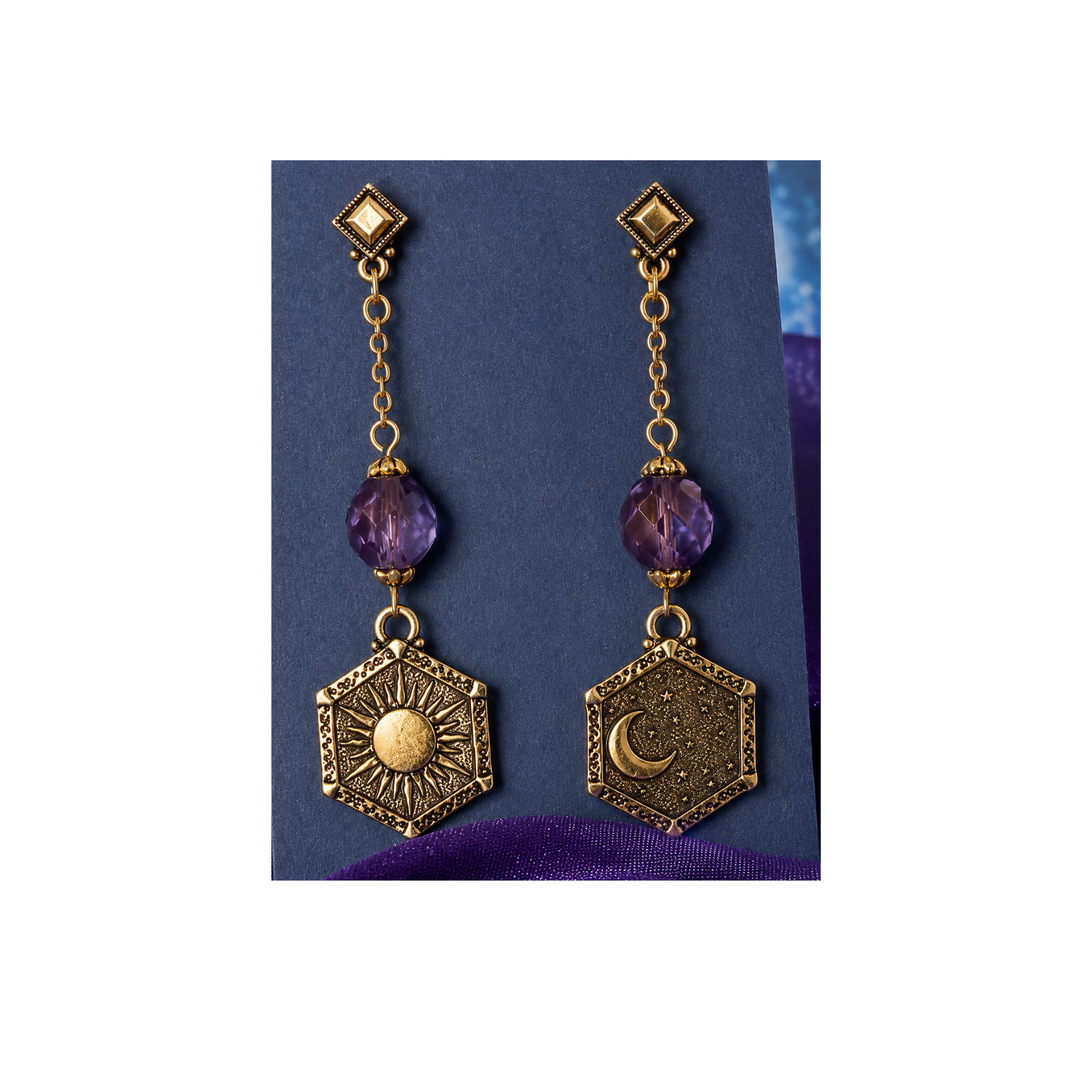 A pair of gold-toned dangle earrings with faceted purple beads and hexagonal charms, one featuring a sun design and the other a crescent moon, displayed on a dark blue card.