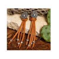 A pair of bohemian-style dangle earrings made from tan leather fringe with silver-toned metal caps and beads, displayed on a rustic background of wood, stone, and a cactus.
